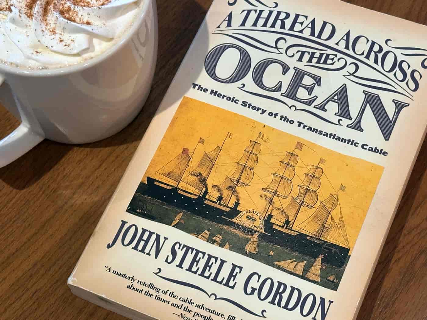 The book 'A Thread Across the Ocean' beside a white mug with whipped cream on a wooden table