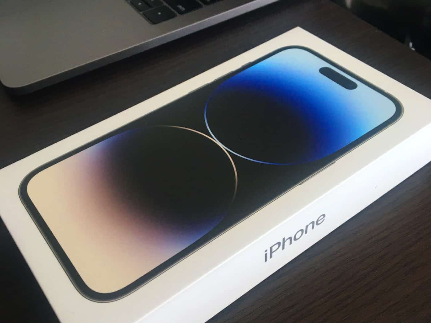 iPhone 14 Pro box with an image of the phone displayed on the front, placed on a wooden desk surface next to a laptop