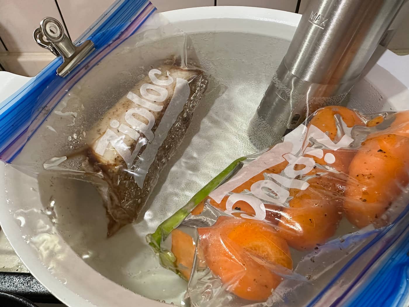 A pot with a sous vide stick heating up water. Steak and carrots in zip-lock plastic bags are immersed for cooking.