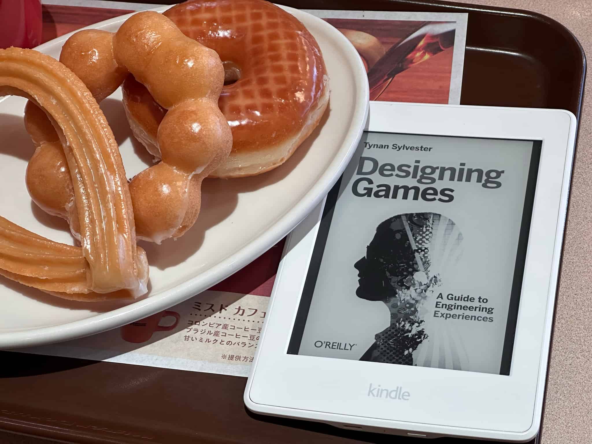 A Kindle e-reader displaying the cover of Designing Games by Tynan Sylvester rests on a tray next to a plate of donuts, including a glazed and a churro, symbolizing a relaxing read with breakfast for game developers looking towards 2024