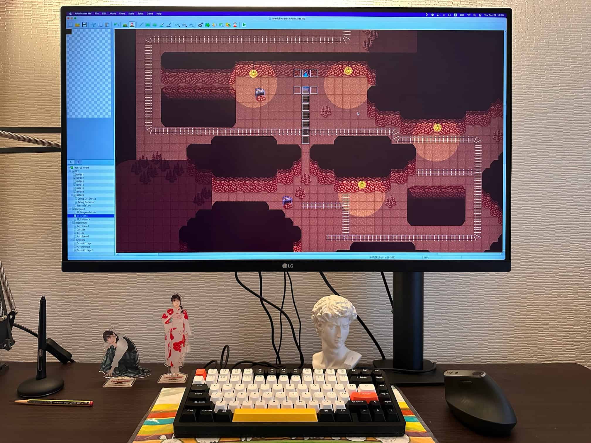 Computer monitor showcasing the RPG Maker MV software interface, with an in-progress game level design. The desk setup includes a mechanical keyboard, a Wacom graphic tablet pen, a vertical mouse, and two acrylic stand figures of Nako Misaki