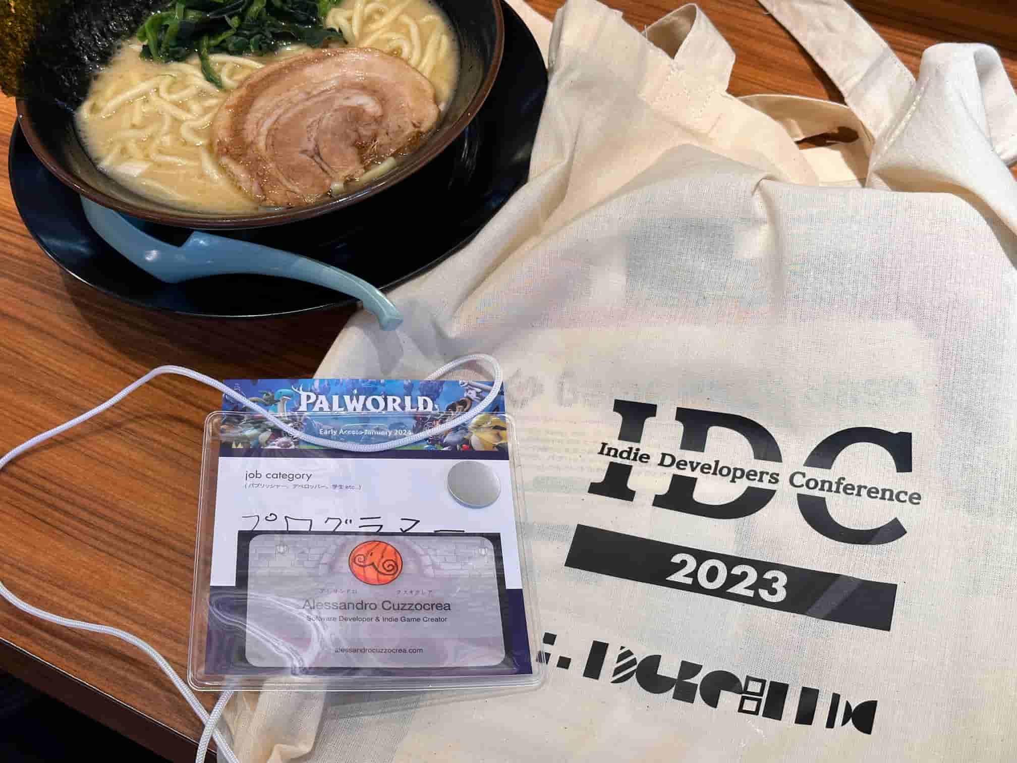 Personal items from the Indie Developers Conference 2023 on a table, including a bowl of ramen, a conference lanyard, and a tote bag with the event's logo. Visible is a badge with the name Alessandro Cuzzocrea, identifying as a Game Developer & Indie Game Creator