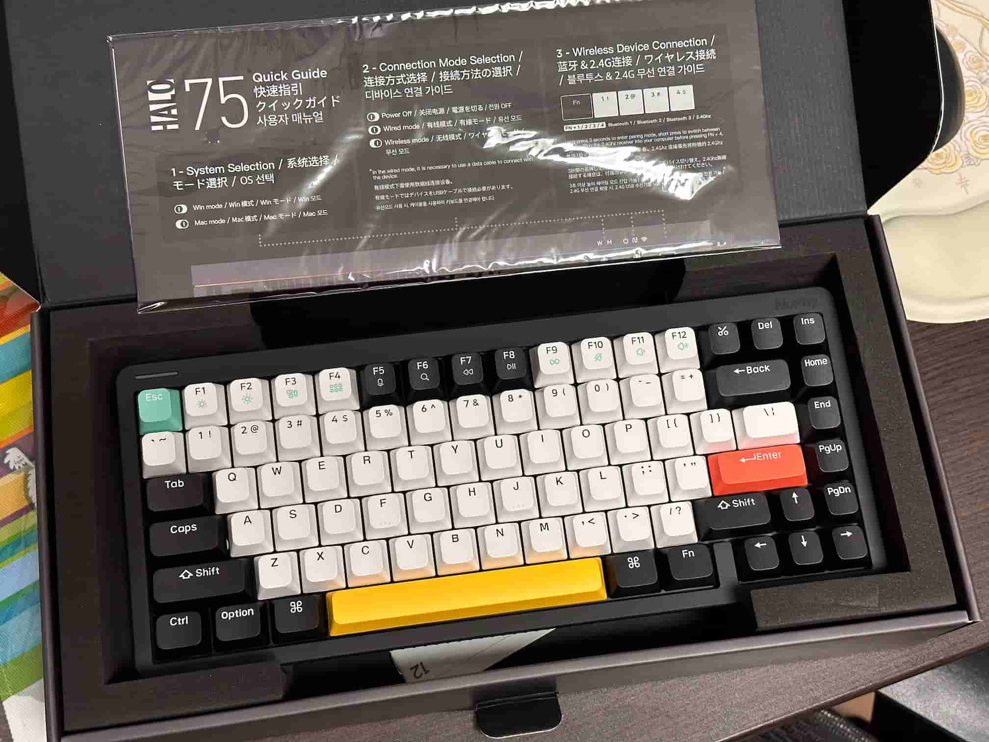 The NuPhy Halo75 mechanical keyboard is showcased inside its box, with the Quick Guide visible on the left. The keyboard features a colorful layout with white, gray, and black keycaps, accented with a blue Esc key, yellow space bar, and red Enter key