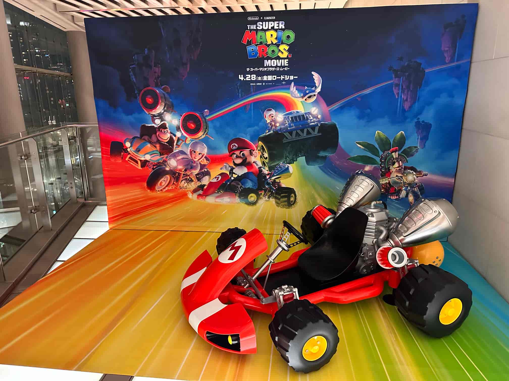 Promotional display for The Super Mario Bros. Movie featuring a life-size model of Mario's red racing kart in front of a vibrant backdrop with characters Mario, Luigi, Princess Peach, and Bowser racing in karts