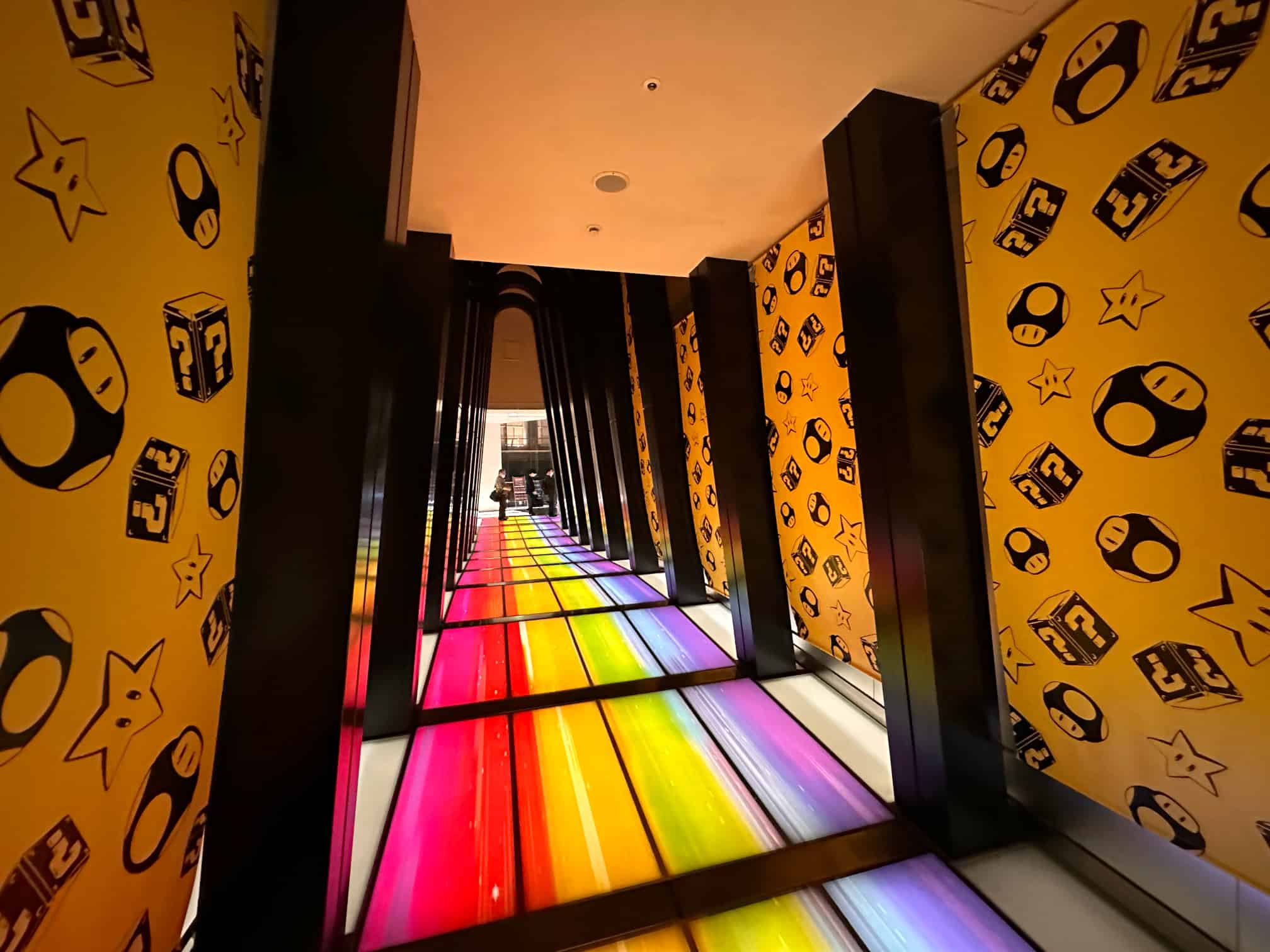 TOHO Cinemas Roppongi Hills corridor inspired by Mario Kart's Rainbow Road, featuring a vibrantly lit floor in rainbow colors and walls adorned with Mario-themed icons such as stars, mushrooms, and question blocks