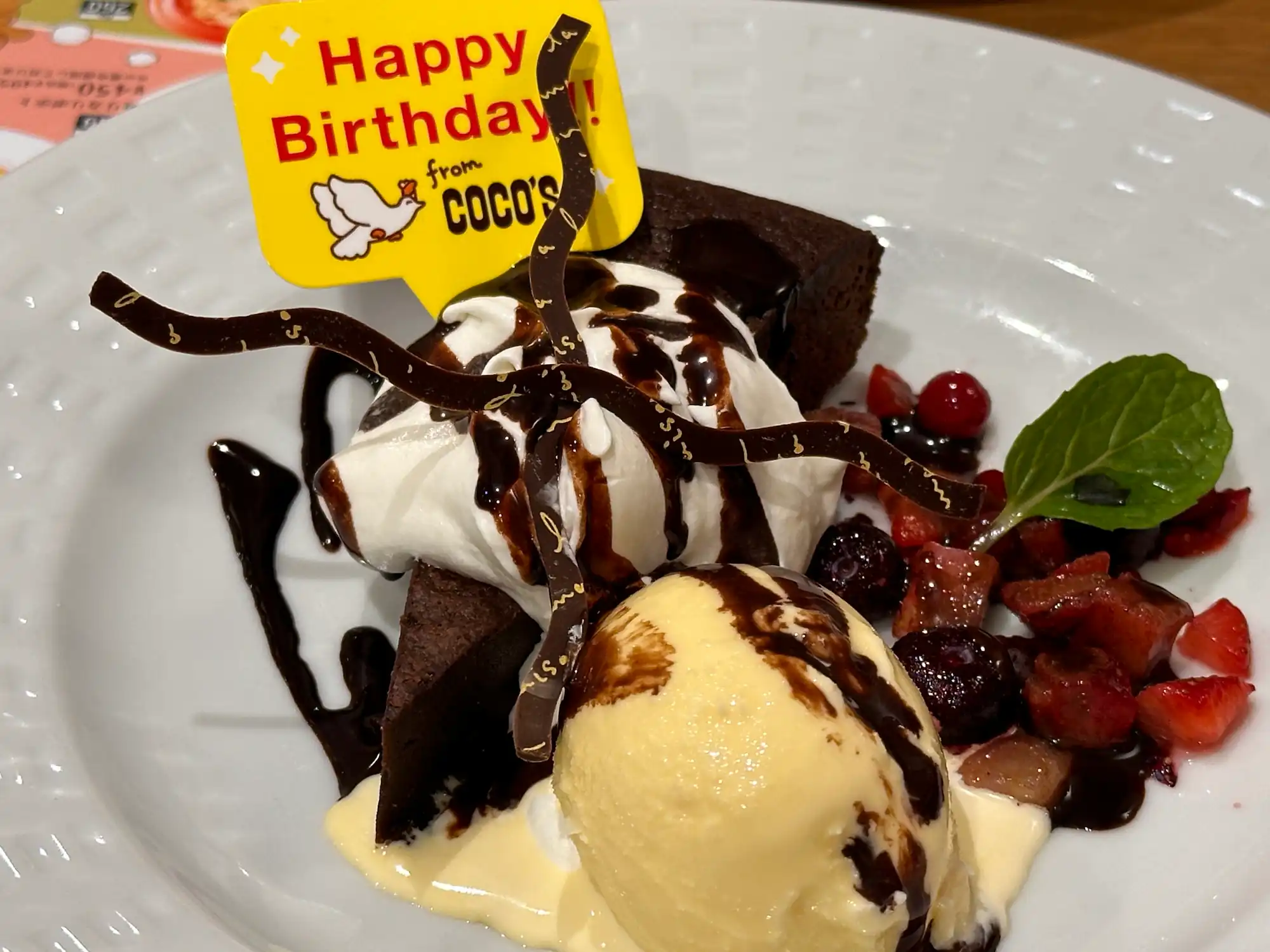 White textured plate holding a slice of chocolate cake topped with whipped cream, decorative chocolate ribbons, and a yellow "Happy Birthday! from COCO'S" sign. The cake is served alongside a scoop of vanilla ice cream, a side of chopped mixed berries with a mint leaf, and is finished with a drizzle of chocolate syrup