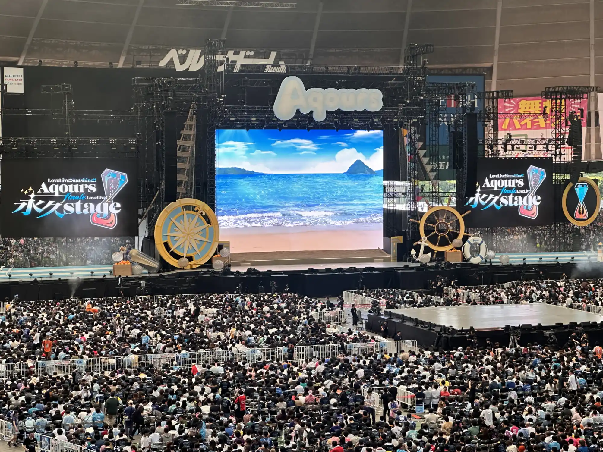 A wide shot of a packed stadium concert for Aqours at Belluna Dome. The elaborate, nautically themed stage features a giant ship's wheel, a compass, and a large central screen displaying a sunny beach, with the official finale LoveLive! logo on two massive side screens