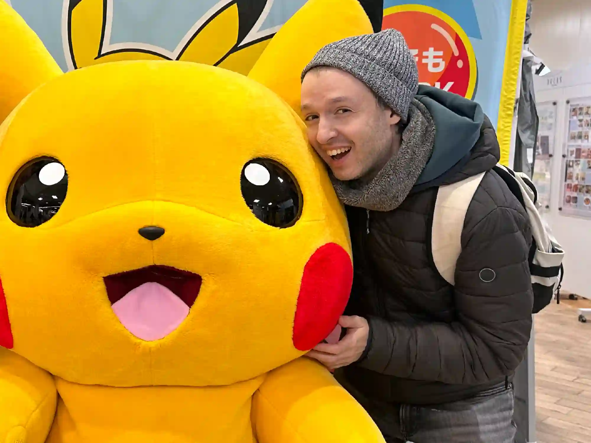 Alessandro Cuzzocrea, smiling and wearing a grey beanie, scarf, and dark puffer jacket, leans cheek-to-cheek against a large, bright yellow Pikachu