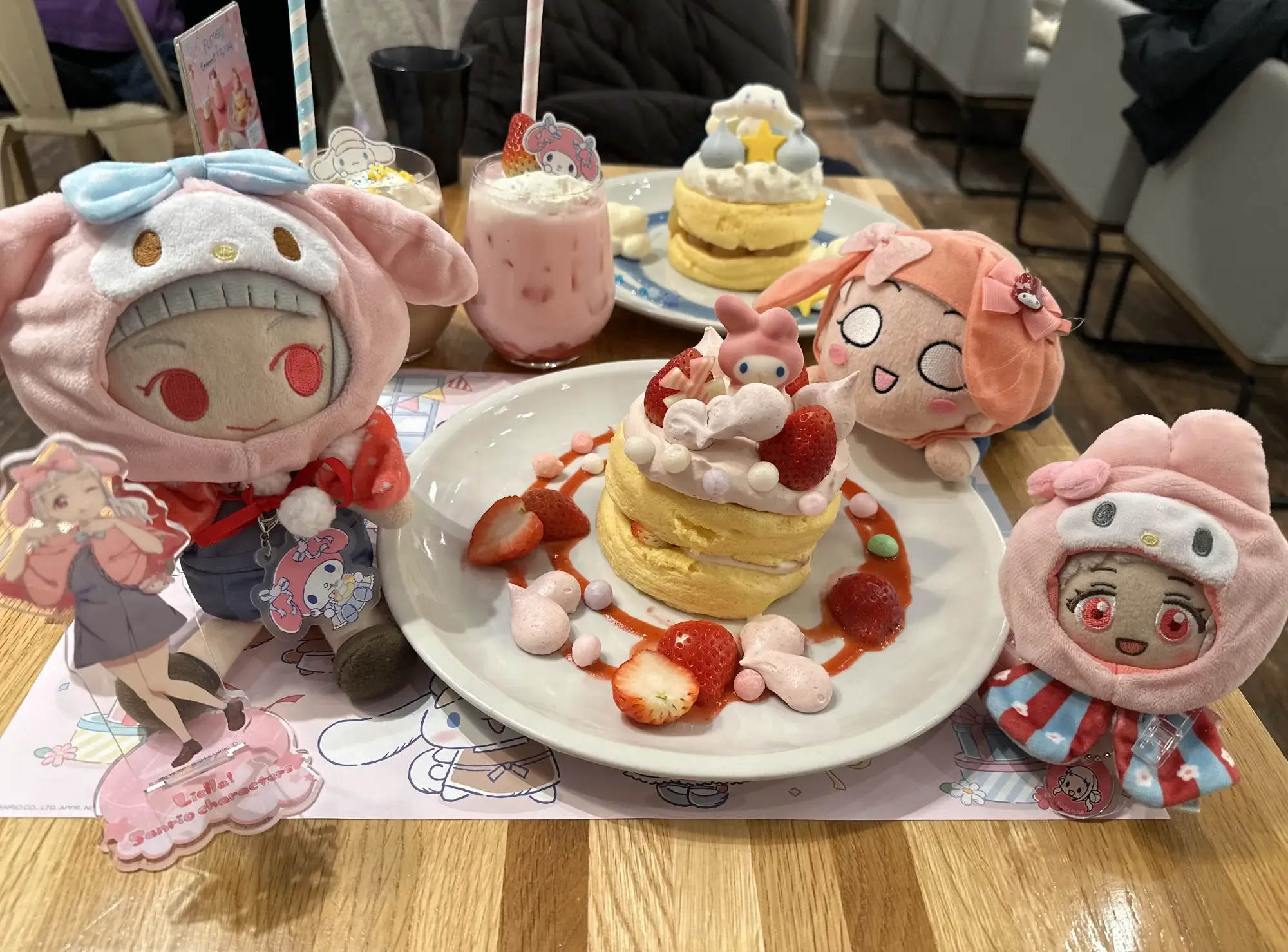 A colorful table at FLIPPER'S Shibuya during a Sanrio collaboration. In the center, a stack of fluffy soufflé pancakes is topped with strawberries and a My Melody figure. The spread is surrounded by pink drinks, a Liella! acrylic stand, and plushies of Chisato Arashi wearing My Melody hoodies