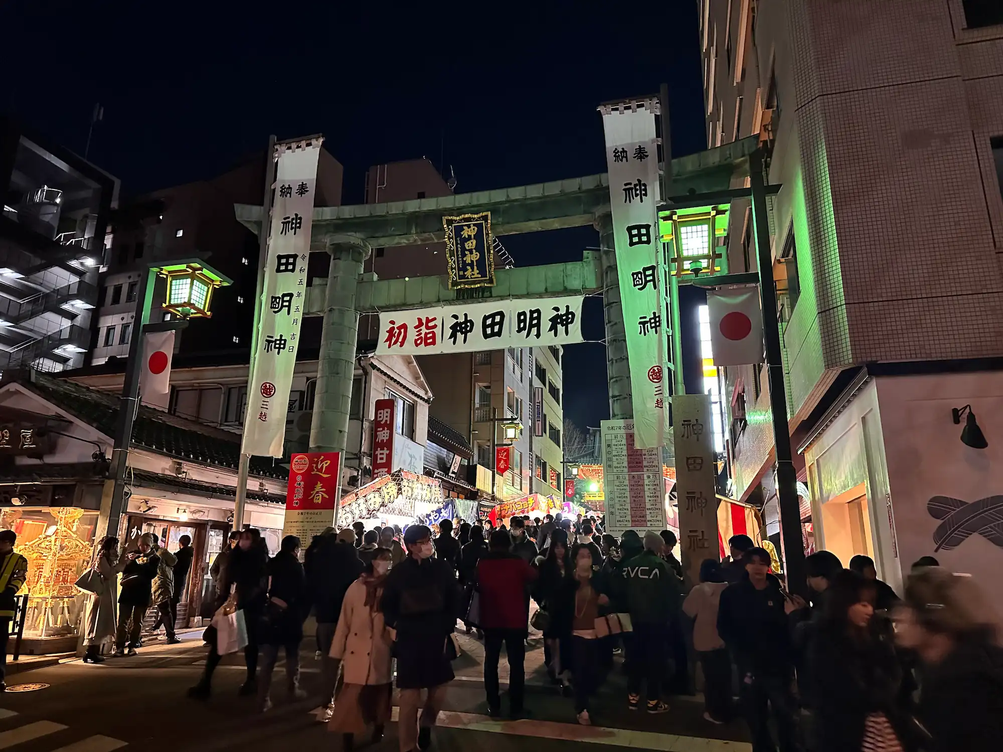 Nighttime view of a dense crowd walking through a large stone torii gate at Kanda Myojin shrine. The gate is flanked by glowing lanterns and Japanese flags, and is decorated with large white banners, including a central one reading "初詣 神田明神" (Hatsumode Kanda Myojin). The brightly illuminated street is lined with buildings and shops as people head toward the shrine area in the background