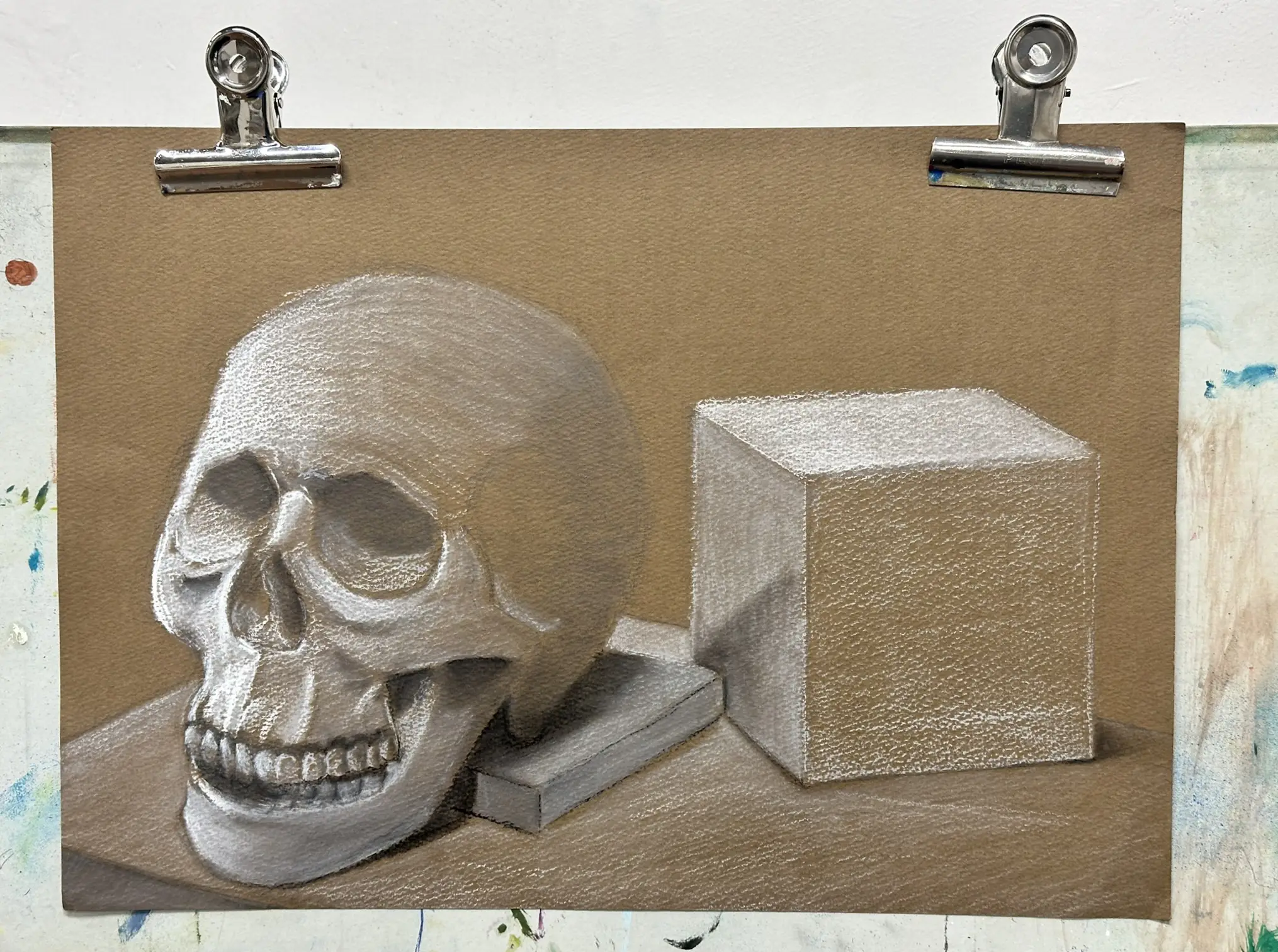 A still life drawing on toned tan paper featuring a human skull on the left and a geometric cube on the right