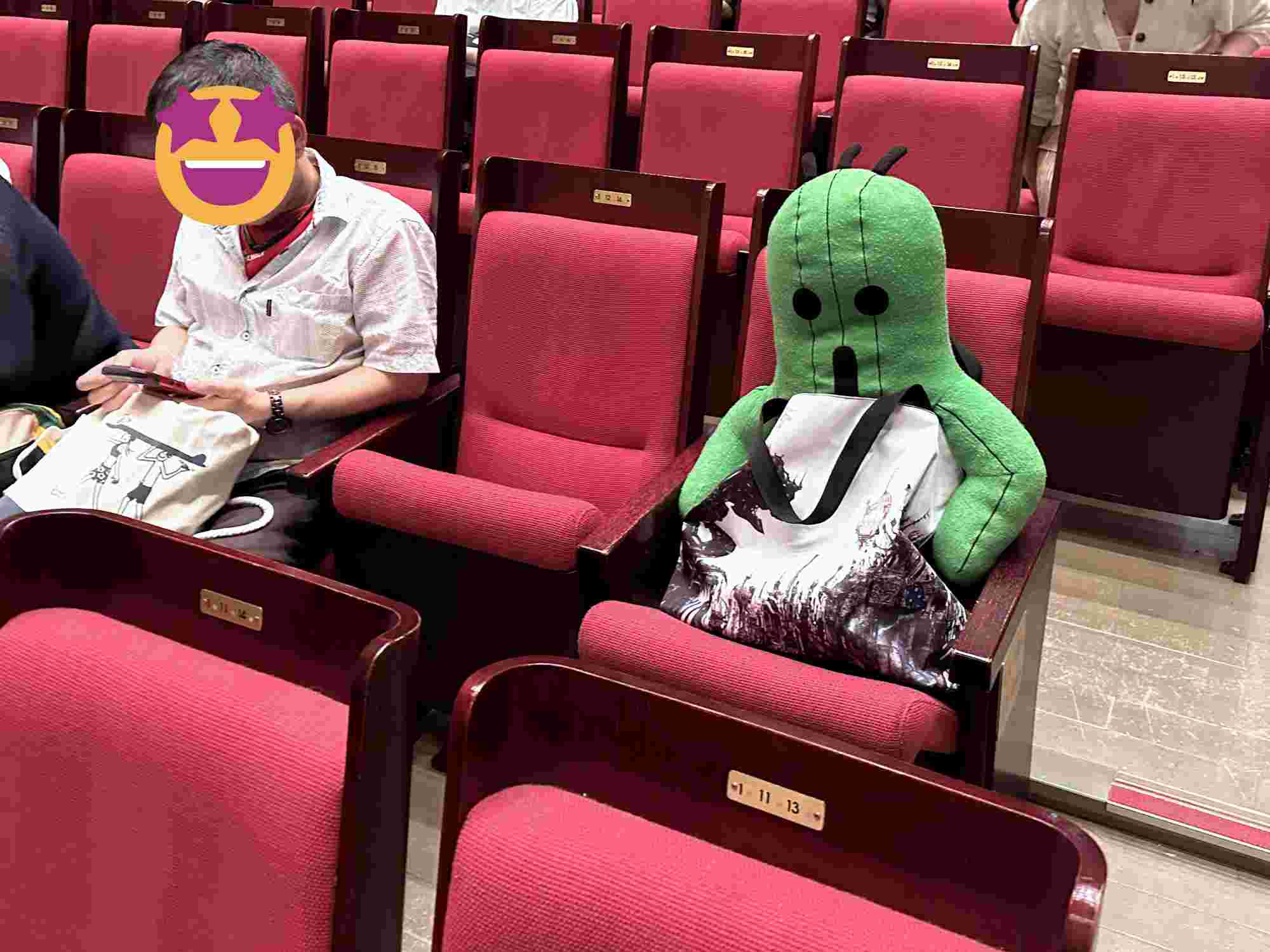 Plush Cactuar toy seated in a concert chair, holding a tote bag, surrounded by red theater seats