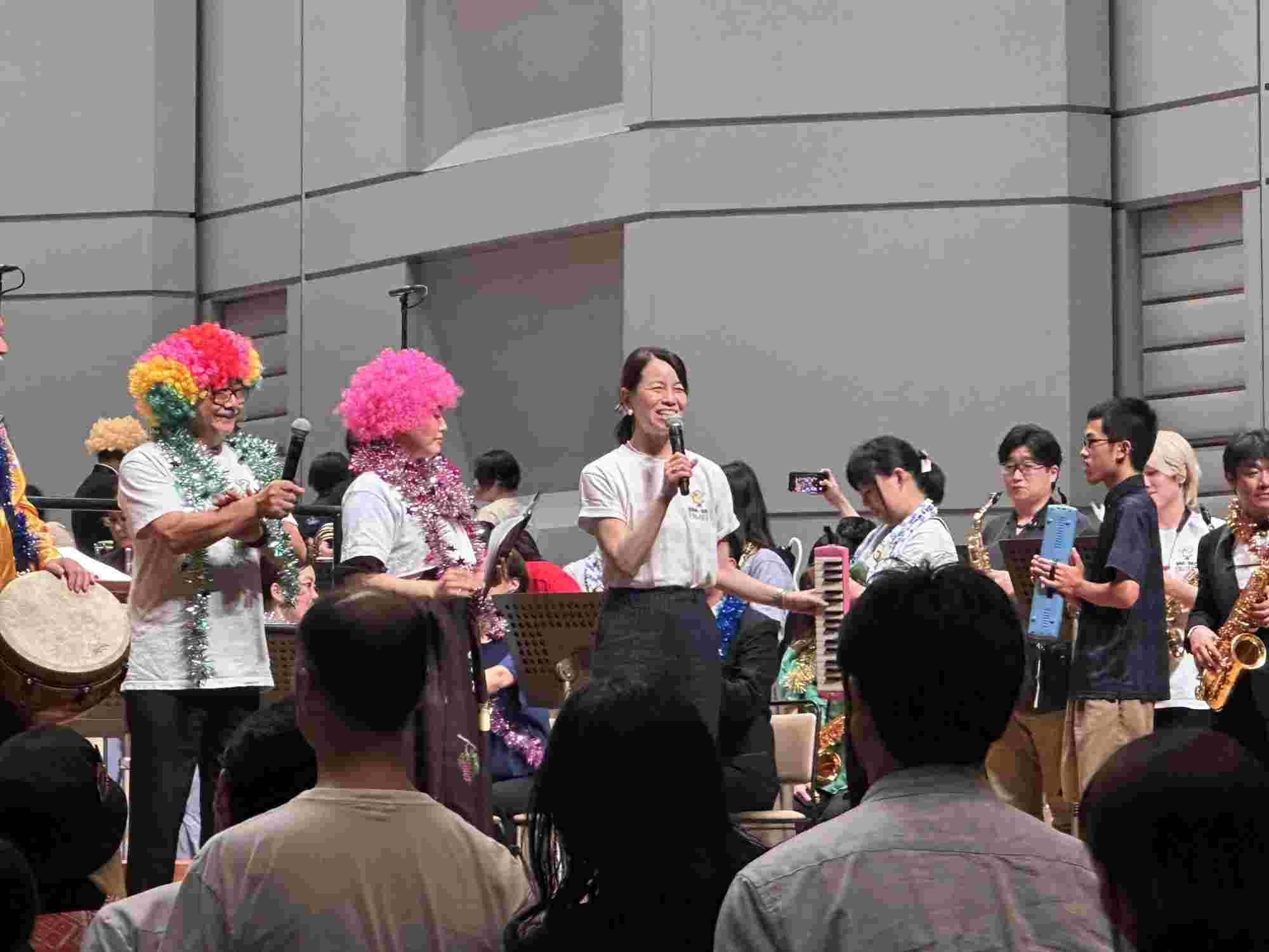 Kazuko Shibuya speaking on stage at the BRA★BRA FINAL FANTASY 2024 concert, alongside Nobuo Uematsu and other musicians in colorful wigs and tinsel