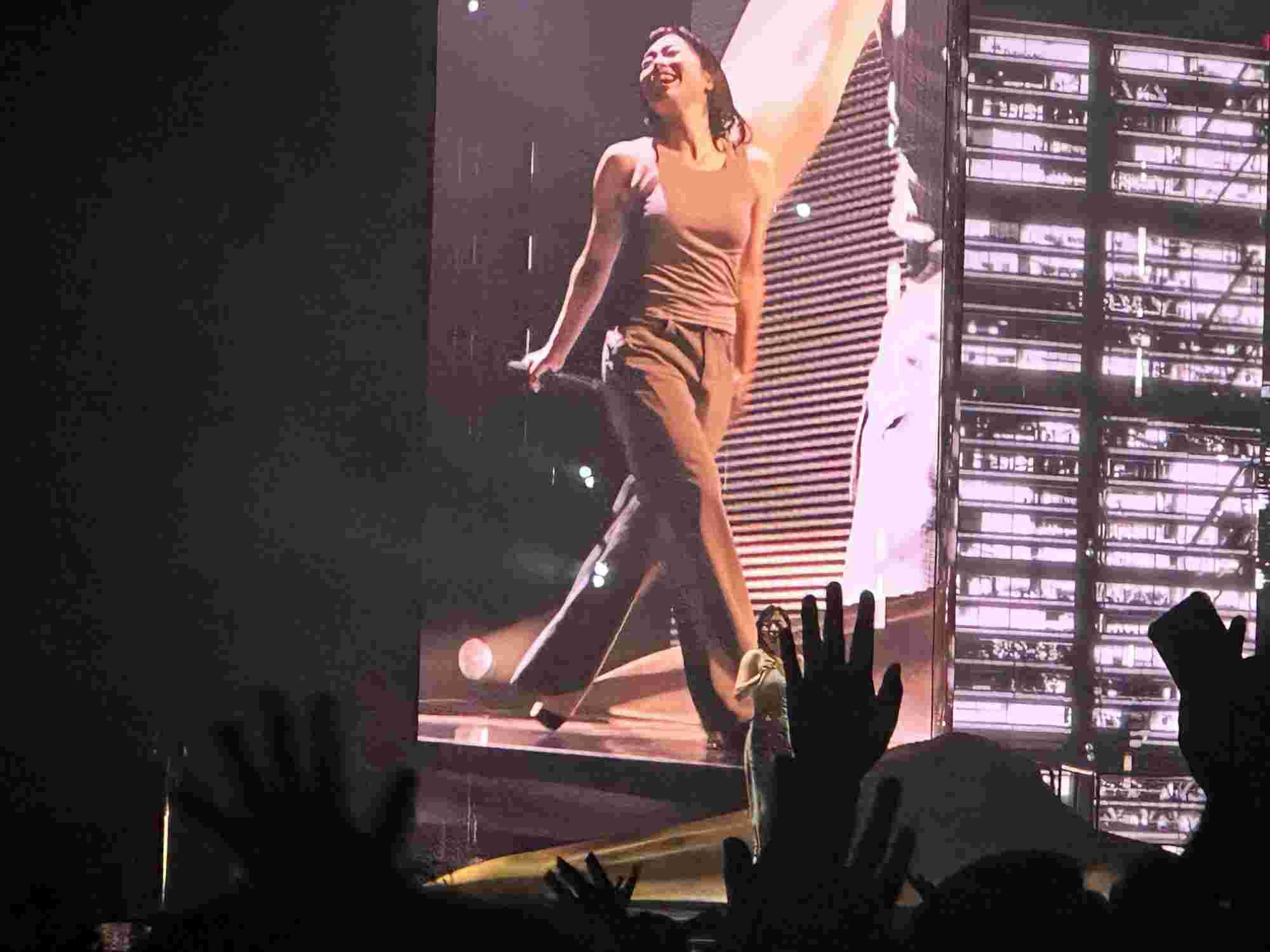 Hikaru Utada smiling brightly while walking across the stage, wearing a relaxed outfit, with their image projected on a large screen in the background. Audience hands are raised in admiration, reflecting the joyful atmosphere of the performance