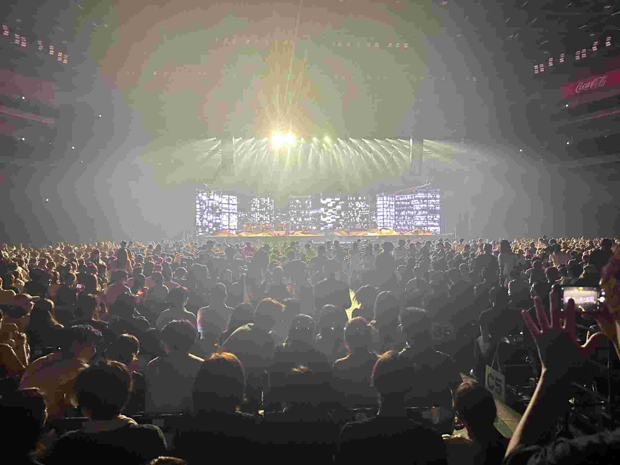 A wide-angle view of a packed concert venue with a brightly lit stage in the distance. The stage features large illuminated screens with dynamic visuals, while the audience fills the foreground, creating a vibrant and energetic atmosphere