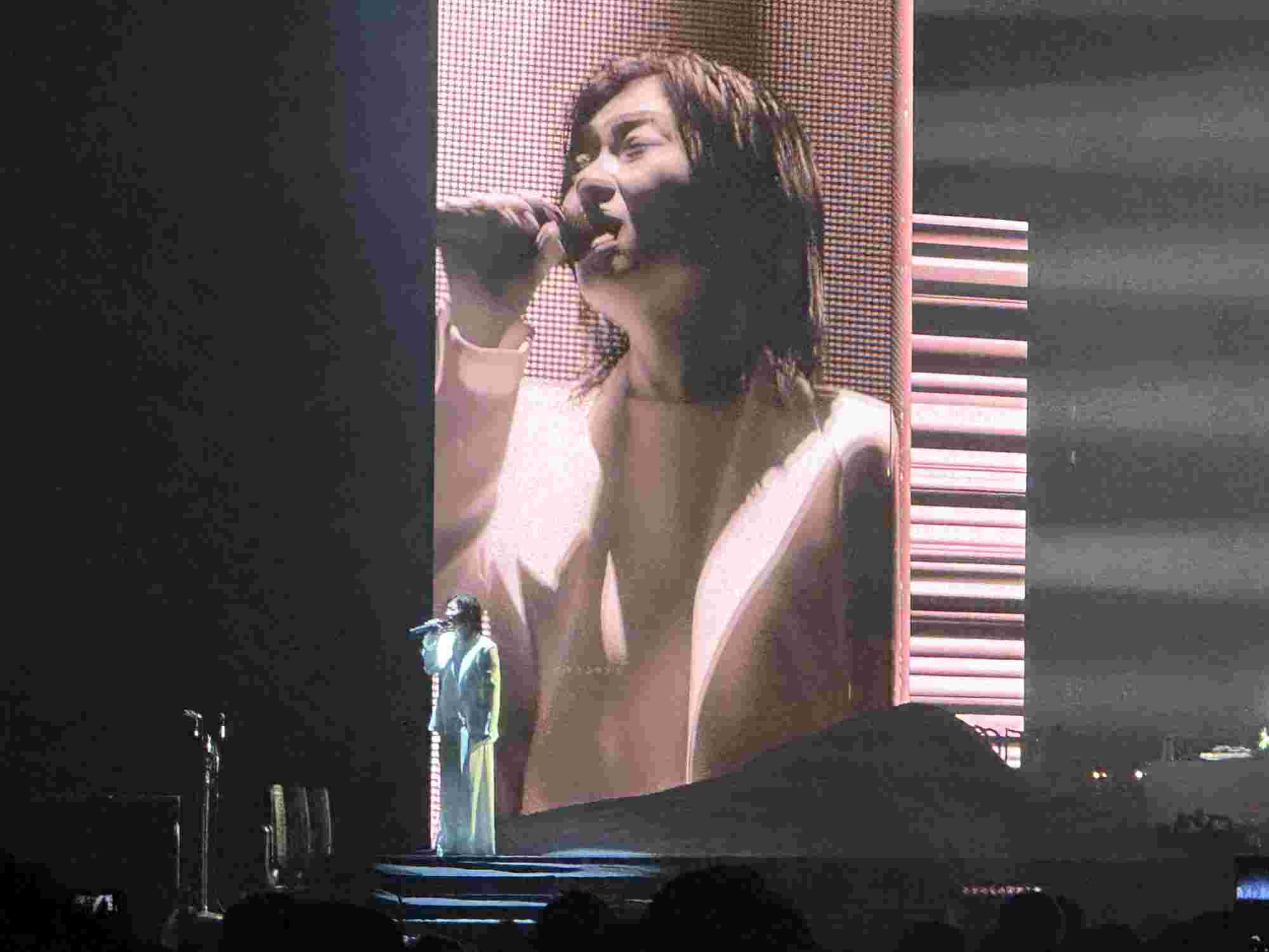 Hikaru Utada performing on stage in a white outfit, singing passionately into a microphone, with a close-up of their face displayed on a large screen behind them