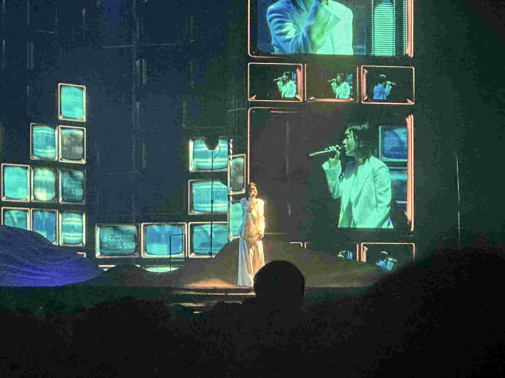 Hikaru Utada performing on stage, dressed in white, surrounded by a backdrop of stacked screens displaying various close-ups of their performance. The glowing blue and green hues from the screens create a retro yet futuristic ambiance as the audience looks on