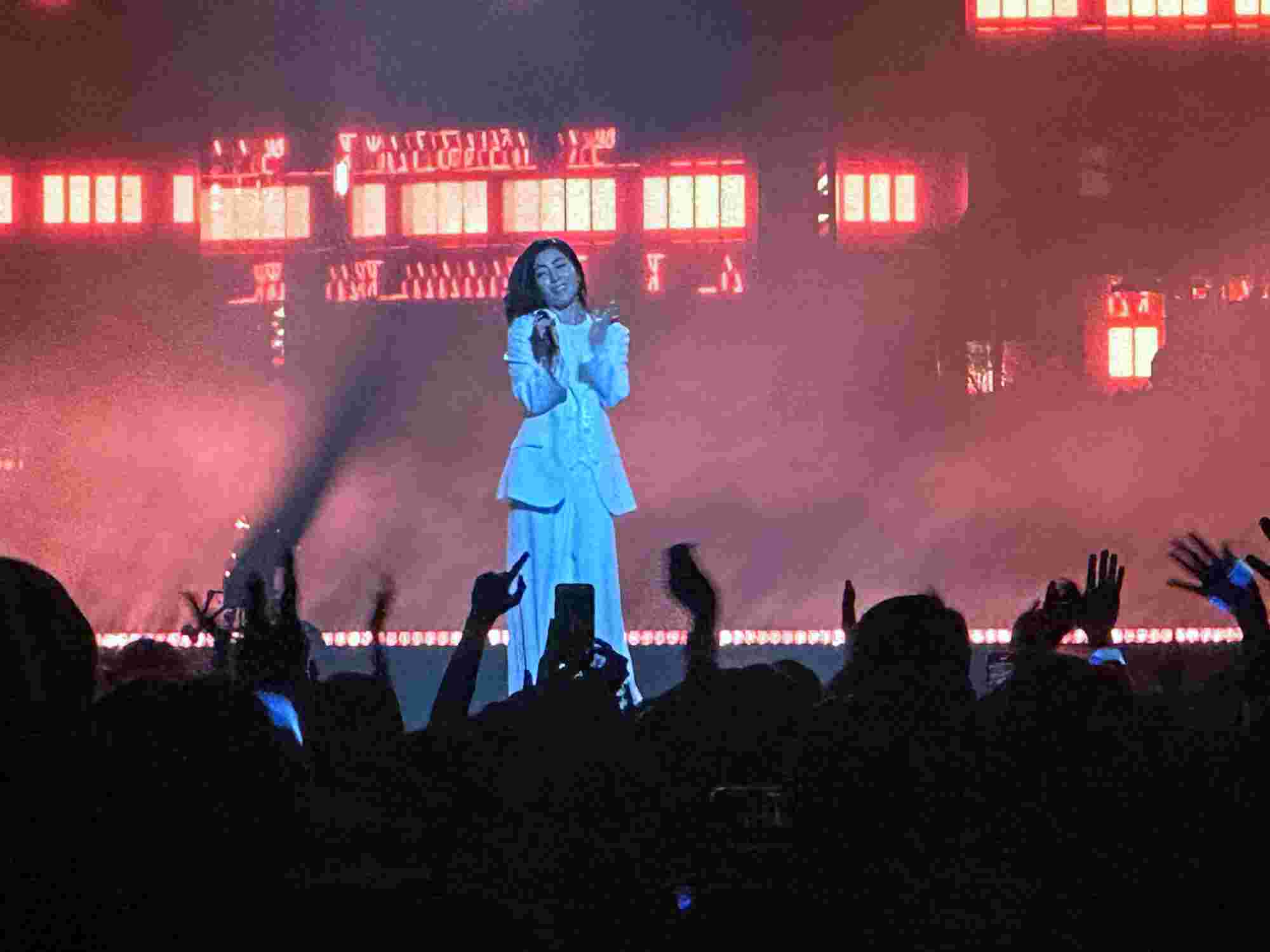 Hikaru Utada on stage, smiling warmly while dressed in a white outfit, surrounded by vibrant red and orange lighting. The crowd in the foreground raises their hands in admiration, contributing to the energetic and emotional atmosphere