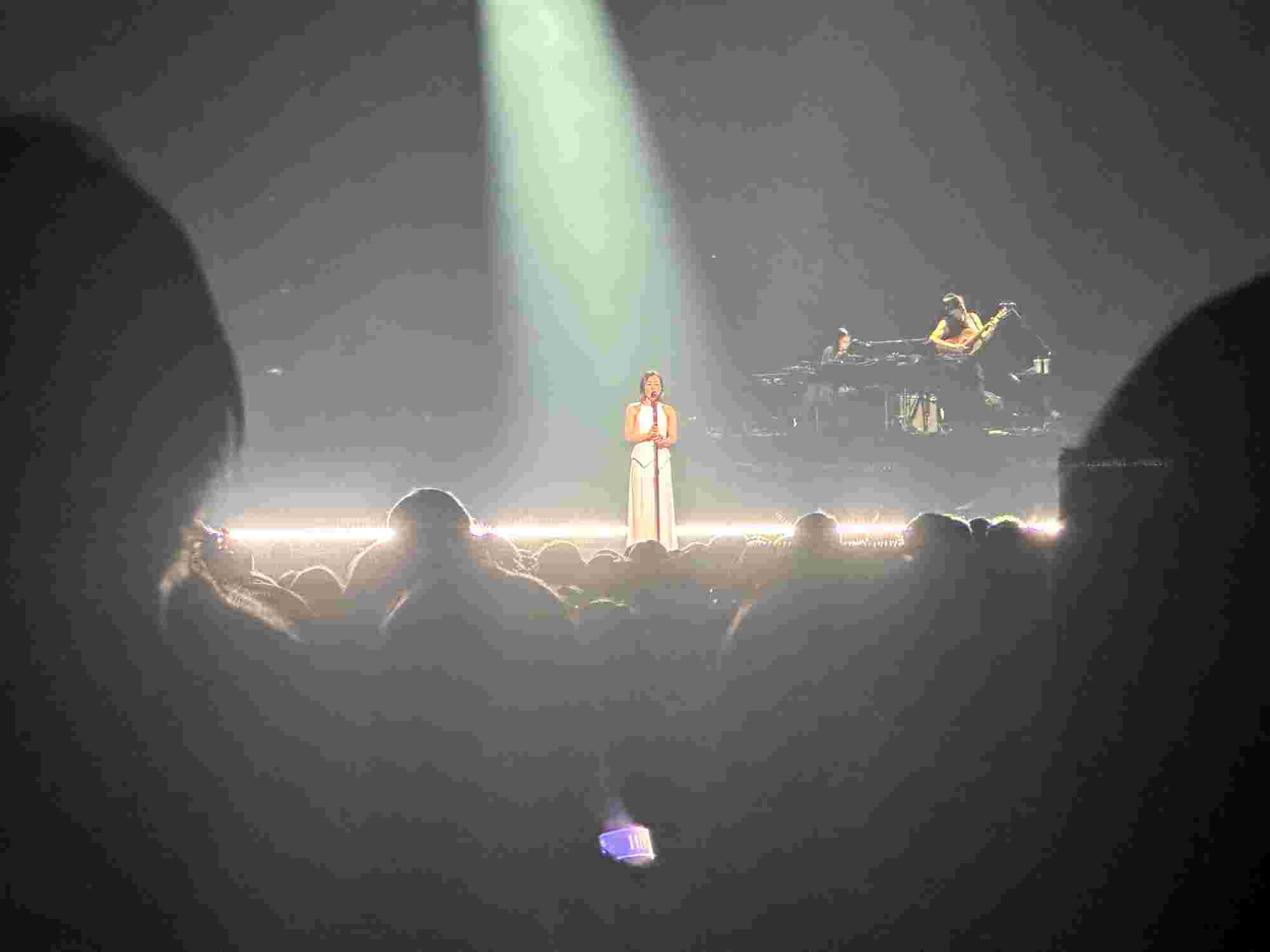 Hikaru Utada stands center stage under a single spotlight, dressed in an elegant white outfit, singing passionately. A live band is visible in the background, adding to the intimate and atmospheric performance, while the audience watches in awe