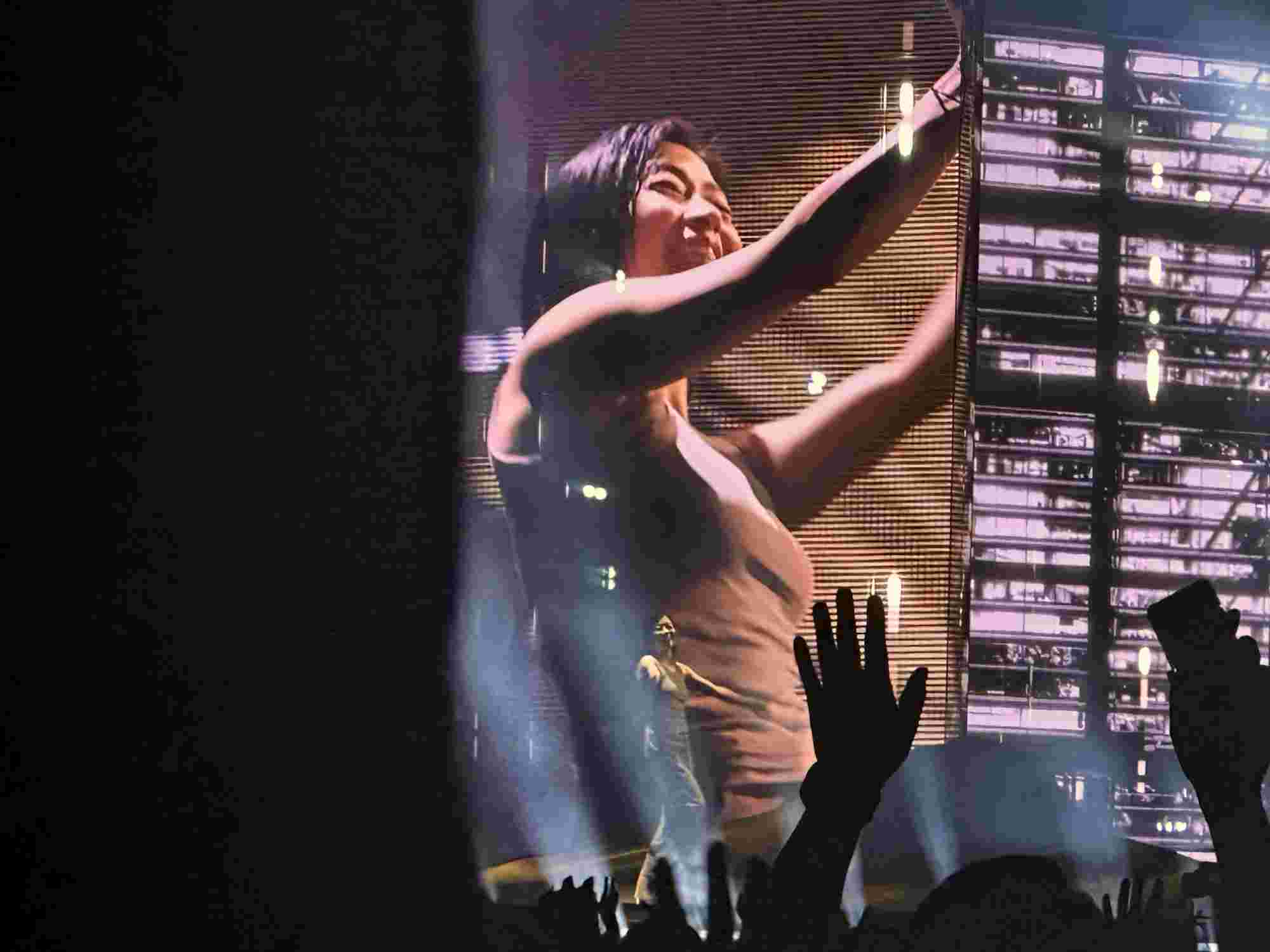 Hikaru Utada joyfully engaging with the audience on stage, raising their arms amidst a backdrop of golden light and an illuminated screen showing their close-up. Hands from the crowd are lifted in response, capturing the energy and connection of the performance