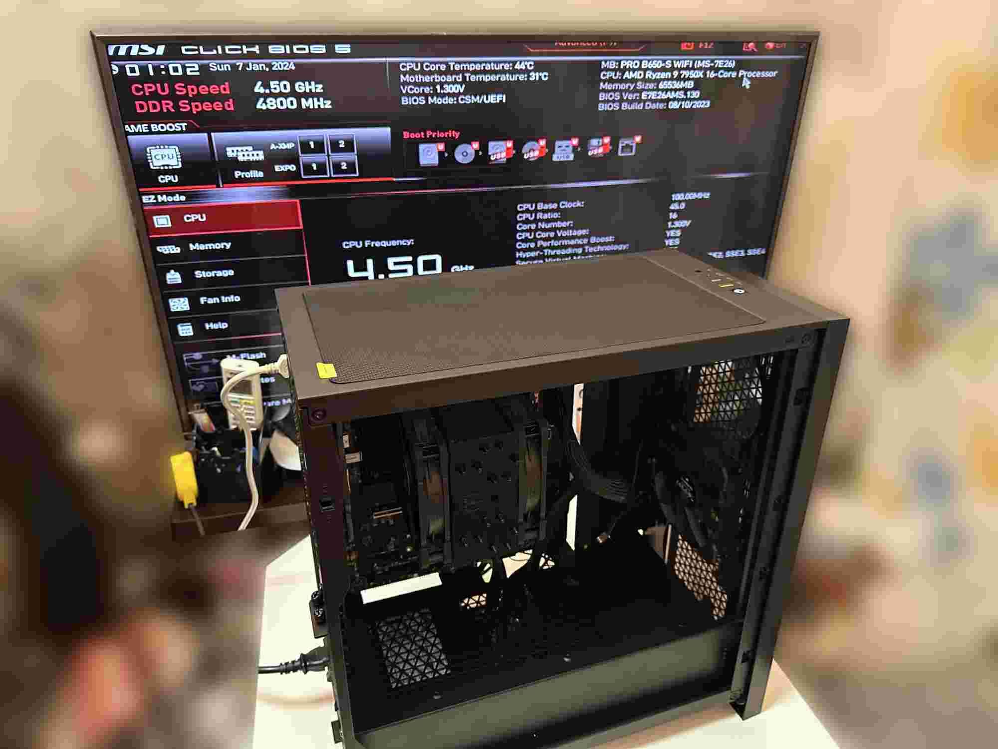 An open black computer case on a table showing internal components, including a CPU cooler and cables. In the background, a monitor displays the MSI BIOS screen with CPU and DDR speed information. The system uses an AMD Ryzen 9 7950X 16-core processor with a clock speed of 4.50 GHz and DDR speed of 4800 MHz