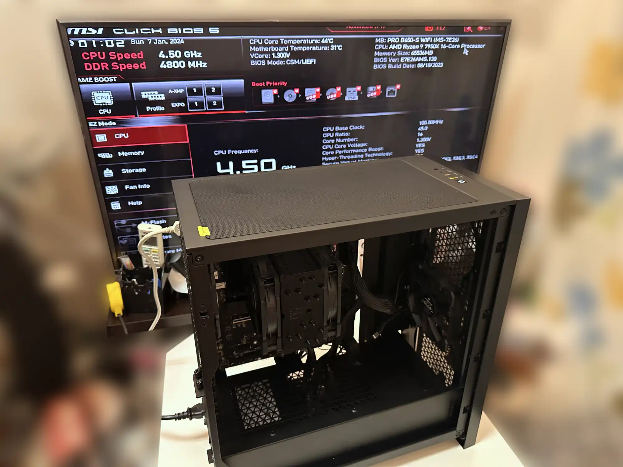 An open black computer case on a table showing internal components, including a CPU cooler and cables. In the background, a monitor displays the MSI BIOS screen with CPU and DDR speed information. The system uses an AMD Ryzen 9 7950X 16-core processor with a clock speed of 4.50 GHz and DDR speed of 4800 MHz