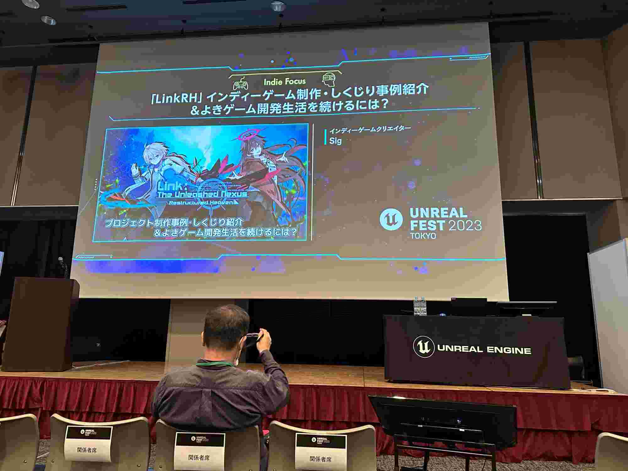 Presentation slide at Unreal Fest Tokyo 2023 titled 'Indie Focus,' showcasing a project called 'LinkRH: The Unleashed Nexus.' The slide features anime-style characters with a futuristic background, alongside text introducing the topic of indie game development and production. The stage setup includes a projector screen, a podium, and a table draped with an Unreal Engine logo cloth, with attendees seated in the foreground